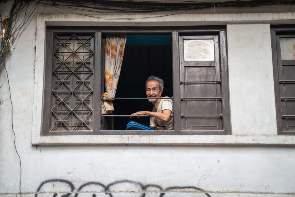 An elderly man in Nepal looks down at the camera from his 2nd floor window.