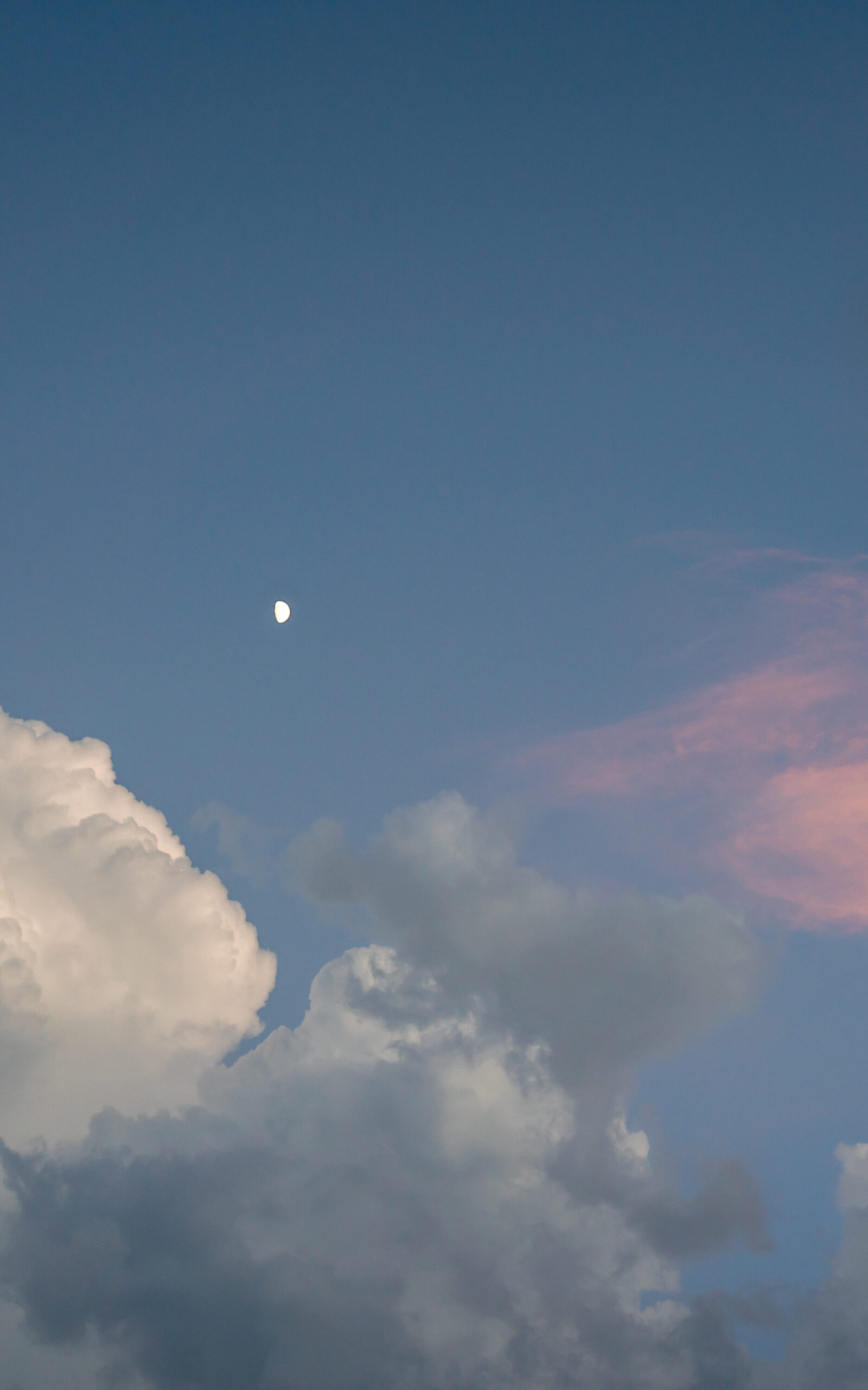 The moon shines brightly in the sky during dusk with white and pink clouds surrounding it.