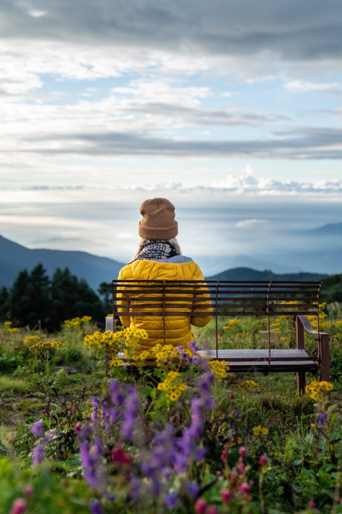 Colorful wildflowers fill the foreground while a woman in a yellow down jacket sits on a bench enjoying the view at the top of Poon Hill in Nepal.