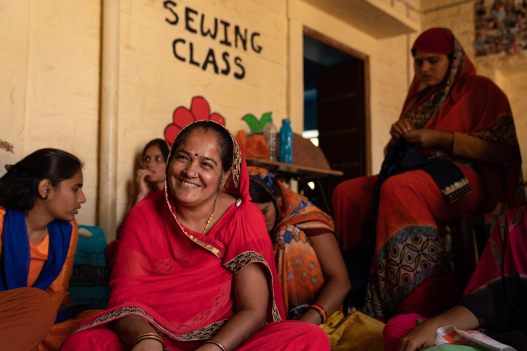 An Indian woman with a red sari on sitting amongst other women in the Sambhali education center in Setrawa, India below a sign that says Sewing Class.