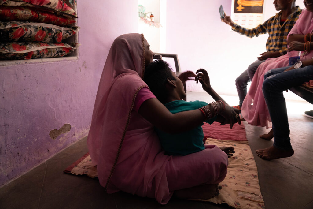 A woman in a pink sari sitting on the floor of her home holding her disabled son.