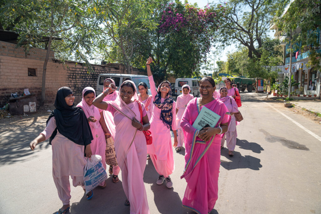 A group of 11 Indian women all in pink saris walking down the street smiling and cheering in unison outside of the Sambhali Trust building in Jodhpur, India.