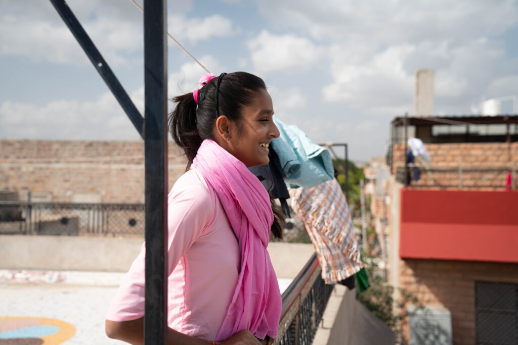 A high school aged girl in a pink sari standing on the top of a boarding home roof looking out into the city with a smile on her face.