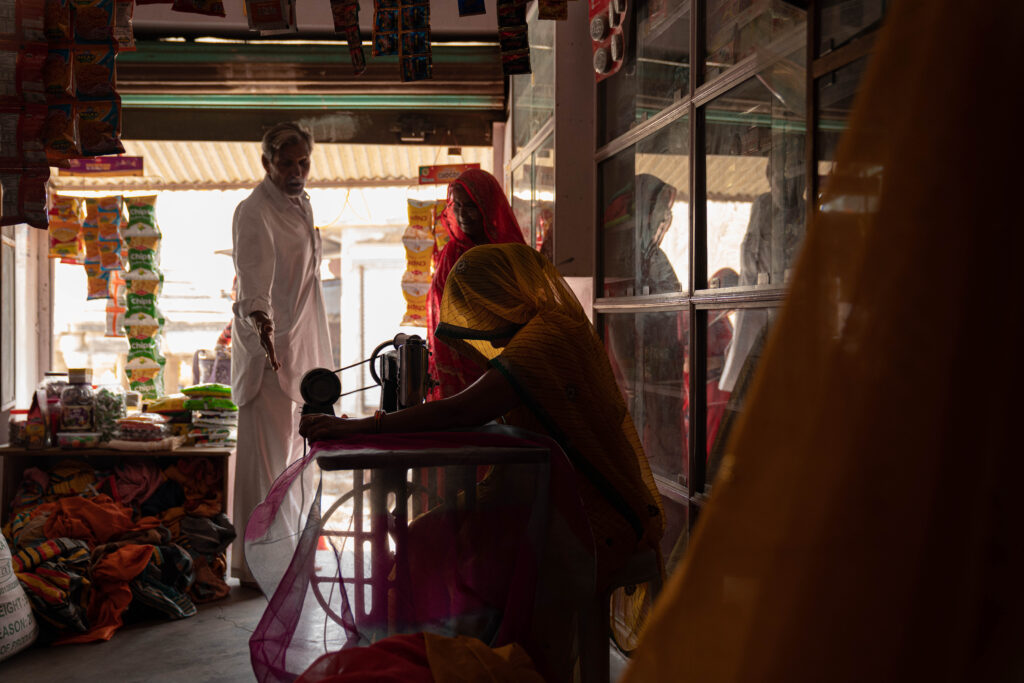 A woman in her shop sewing a sari with a man looking over her pointing down at the floor.