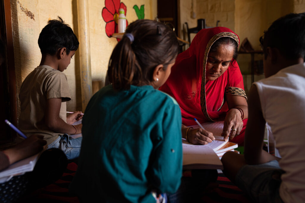 A woman in a red sari tutoring elementary aged children inside the Sambhali education center in the Thar Desert in India.