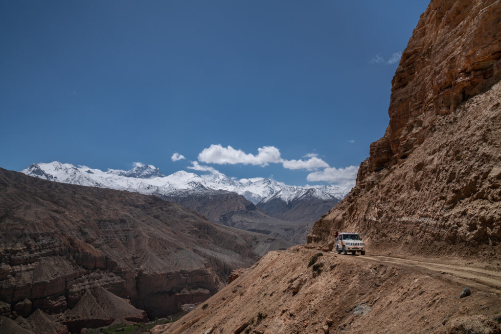 A Mahindra truck drives on a dirt road with a sheer drop of in the Upper Mustang in Nepal with views of snow-capped Himalayan mountains in the background.