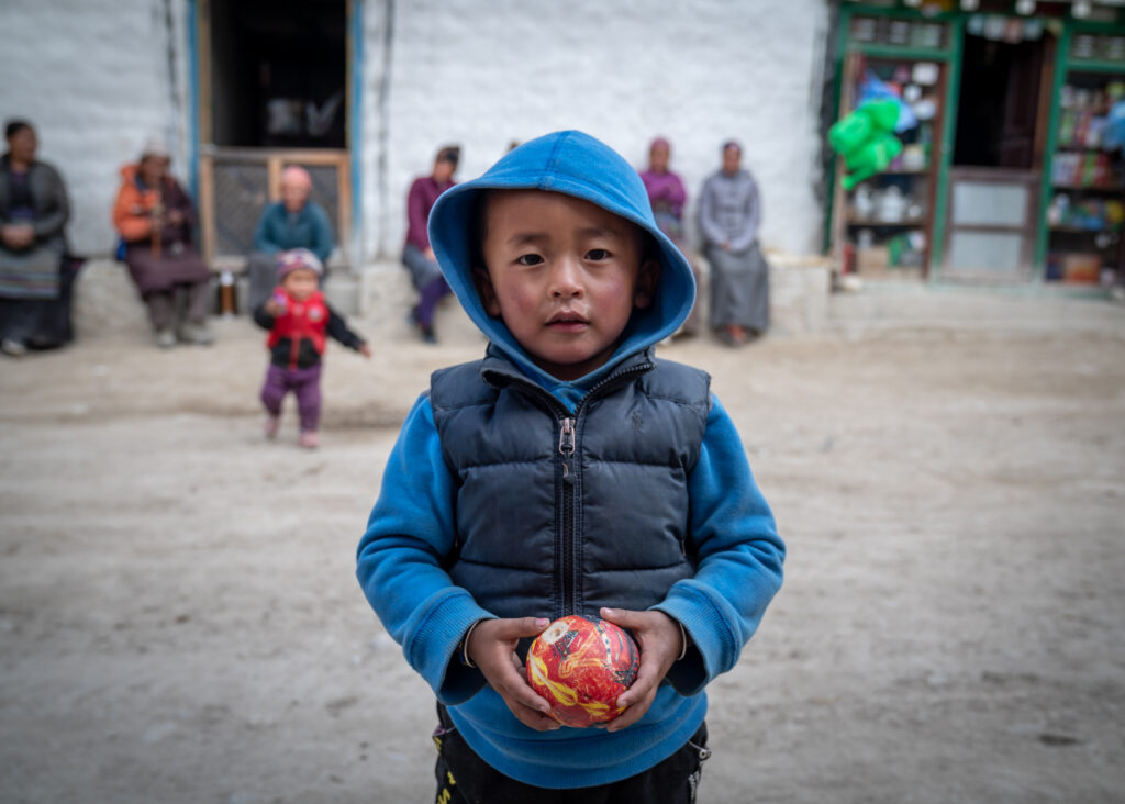 A toddler boy in a blue hoody holding a red and yellow ball in a dirt street in Lo Manthang, Nepal in the Upper Mustang.