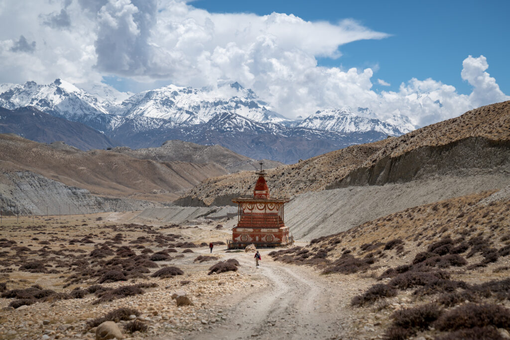 Two people hike down a long dirt road towards a Buddhist Stupa in the middle of nowhere with the snowy Himalayas towering behind it.