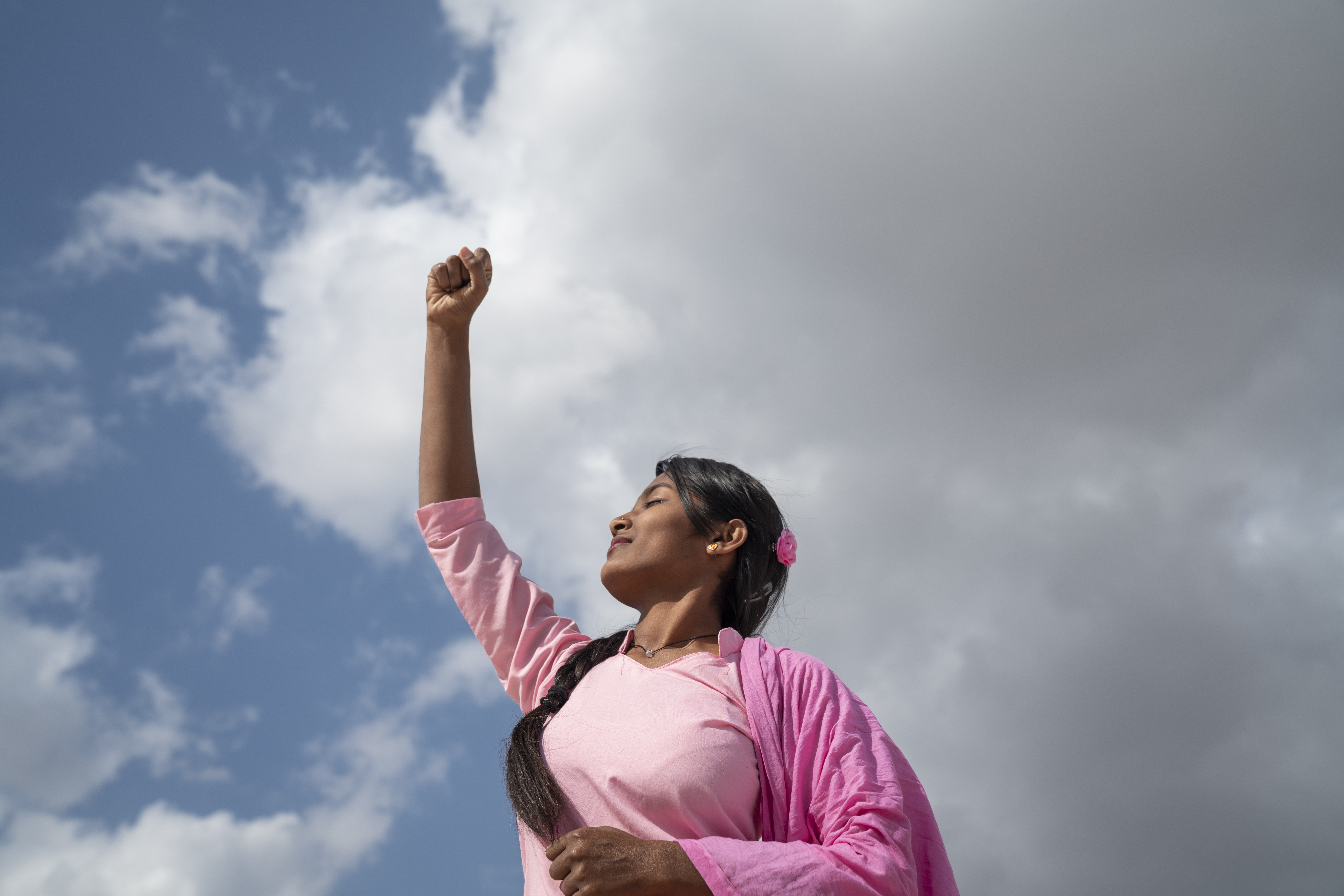 A young Indian woman in a pink outfit empowered with her right hand in a fist raised high in the air with the sky behind her.