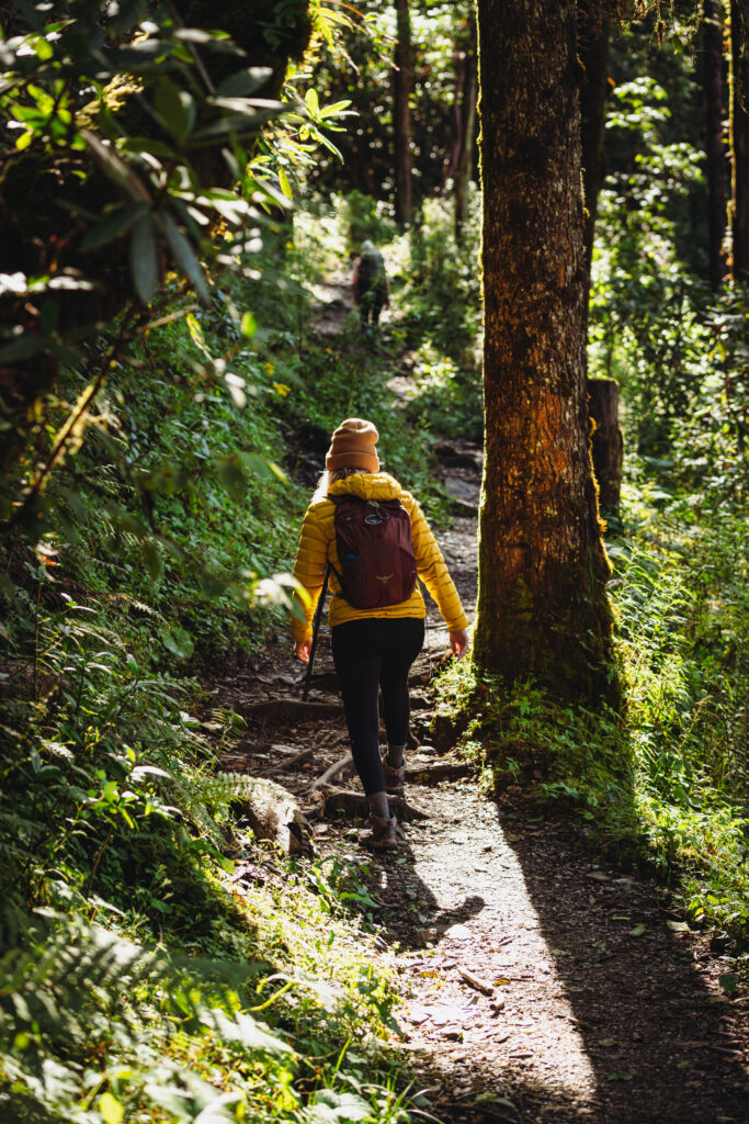 A woman in a yellow coat hiking in a lush forest in Nepal wearing an Osprey daypack.