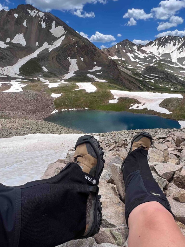 A hikers feet show Yaktrax microspikes and Outdoor Research crocodile gore-tex gaiters with a snowfield, glacier lake and snowy mountains in the background. 