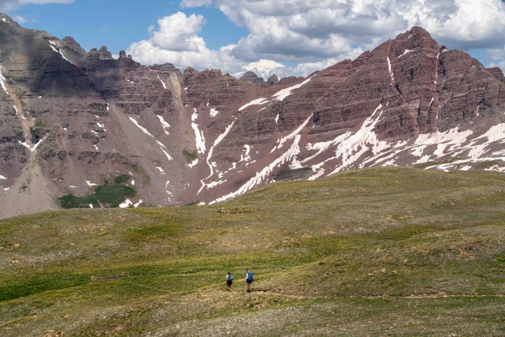 Two women are hiking on a trail on the way to Conundrum Hot Springs in Colorado with red peaks in the background.