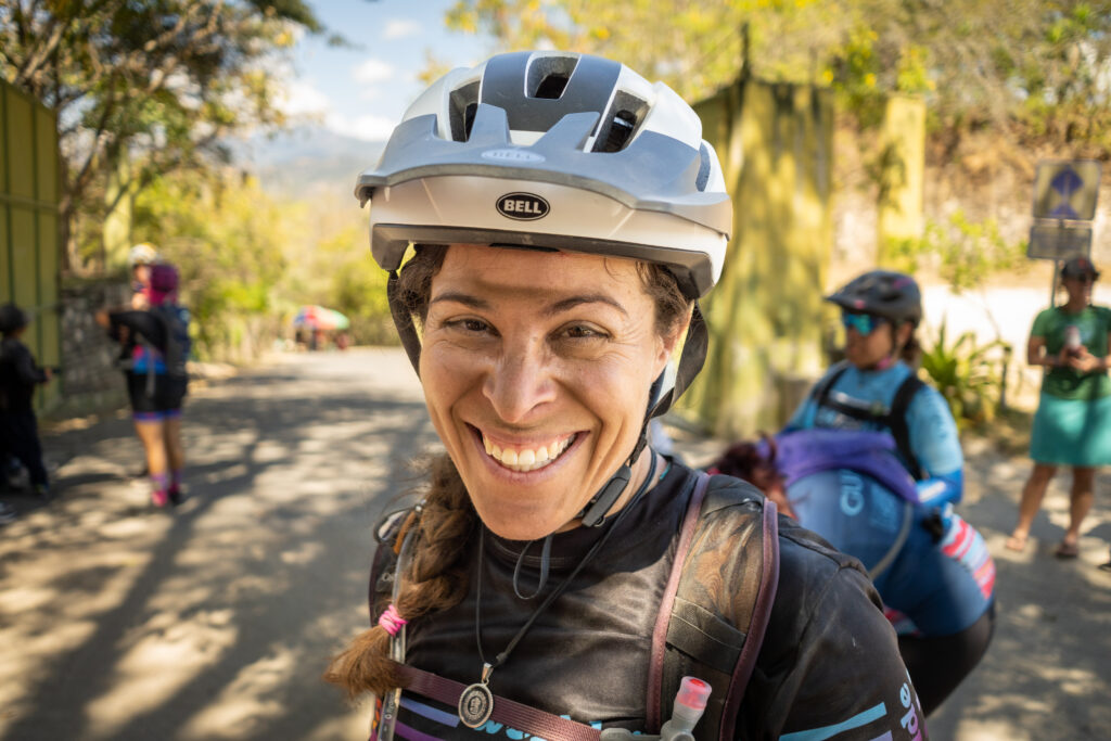 A woman smiles with a dirt unibrow and a bell helmet on her head at the end of a big mountain bike race.
