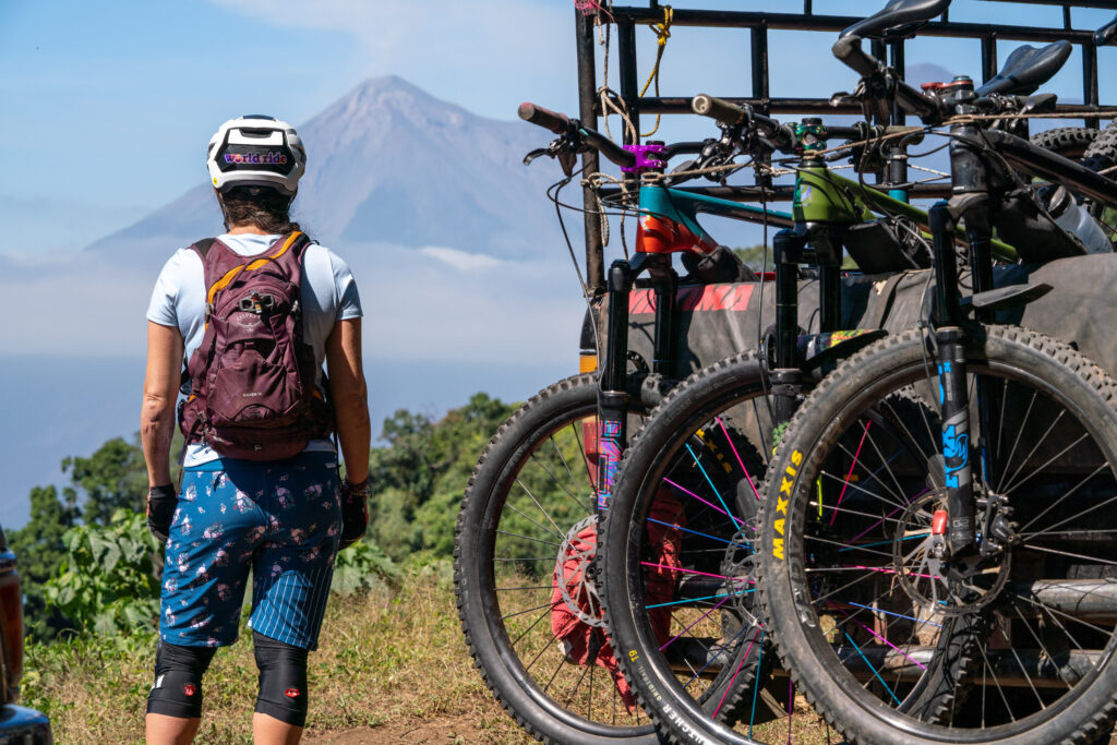 The executive director of World Ride, Julie Cornelius, stands at the top of a viewpoint looking out at El Fuego volcano in Guatemala. There is a truck of 3 mountain bikes next to her.