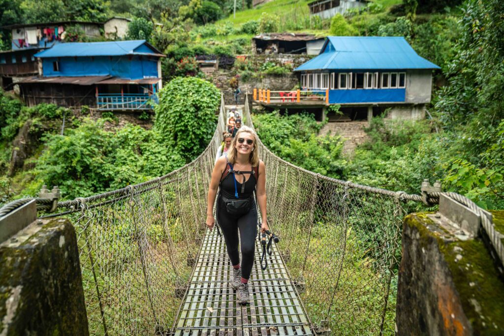 A blonde woman walks across a suspension bridge with trekking poles in hand after completing the Poon Hill Trek in Nepal.