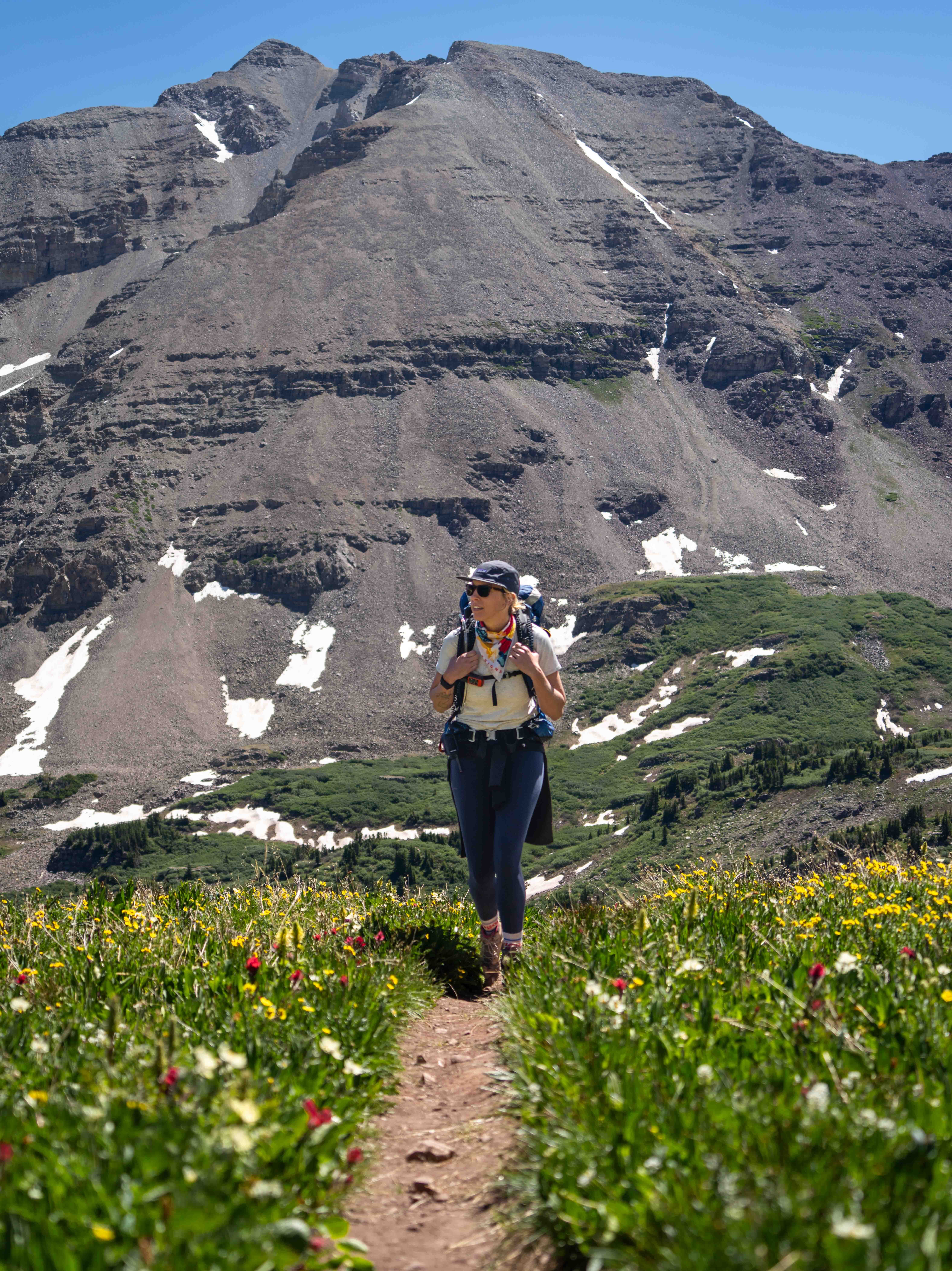 Hilary Lex hikes up a trail surrounded by wildflowers on the Conundrum Creek Trail in Colorado. A mountain towers behind her.