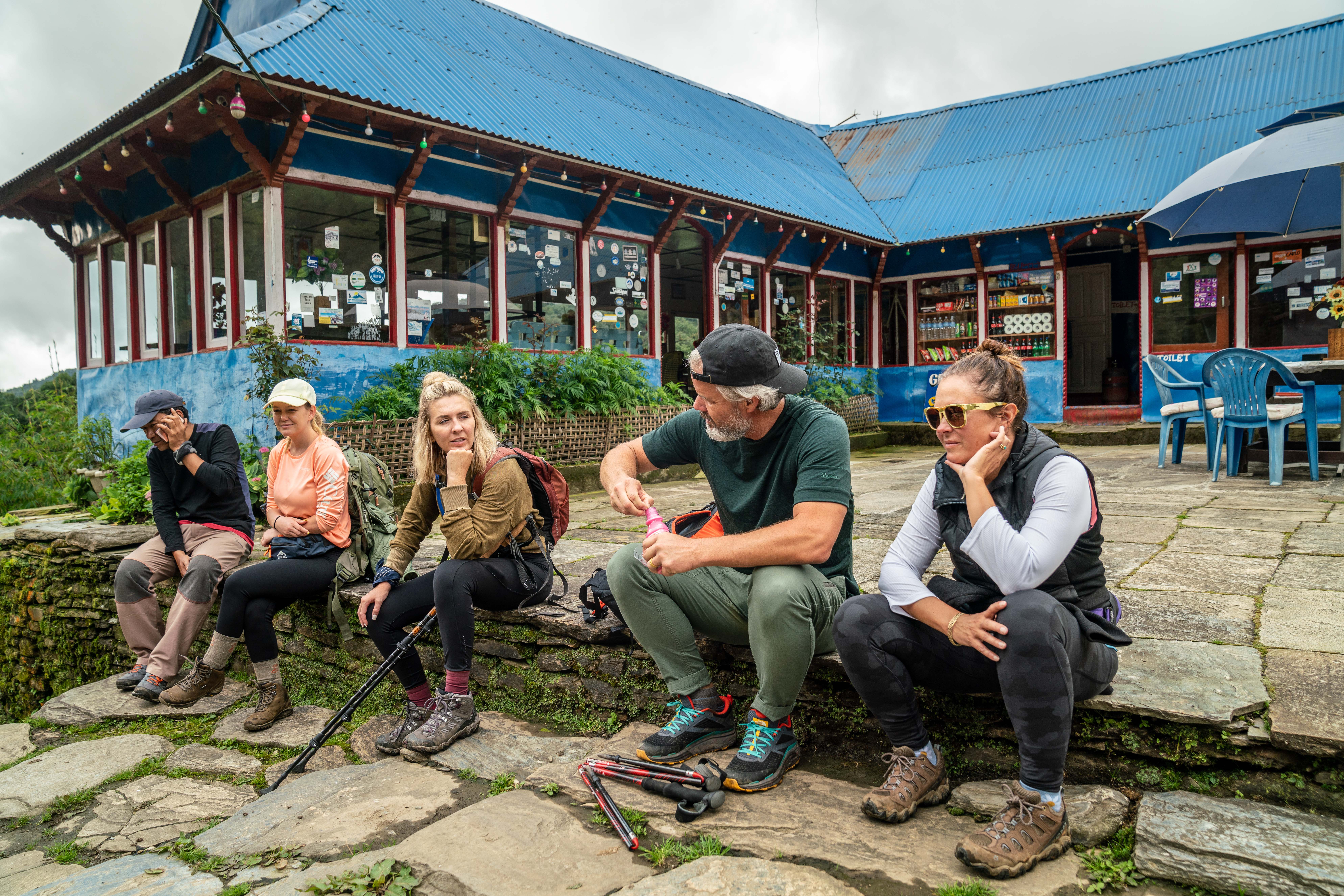 a group of hikers taking a rest break in front of a teahouse in nepal. one hiker is opening a bottle of peptobismal.